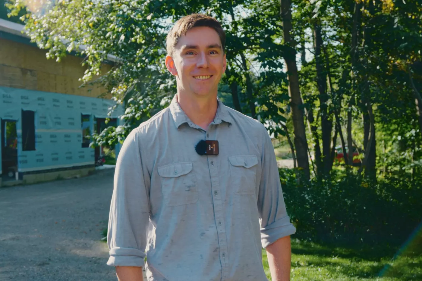 Smiling man in a light shirt stands outdoors, surrounded by trees and construction materials.